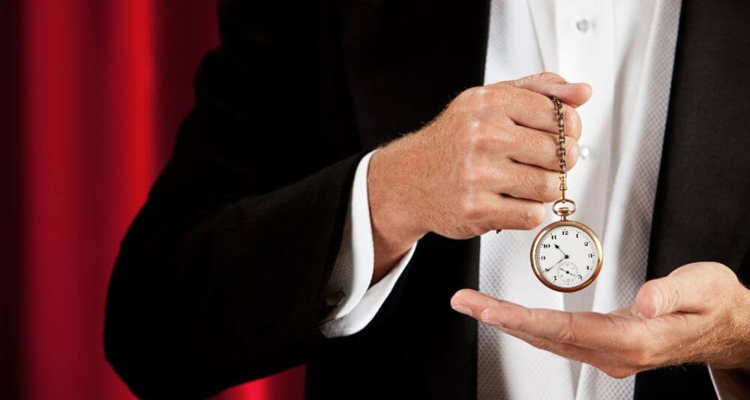 A hand holds a gold pocket watch by its chain against a red curtain backdrop, while another hand is open below, as if catching it.